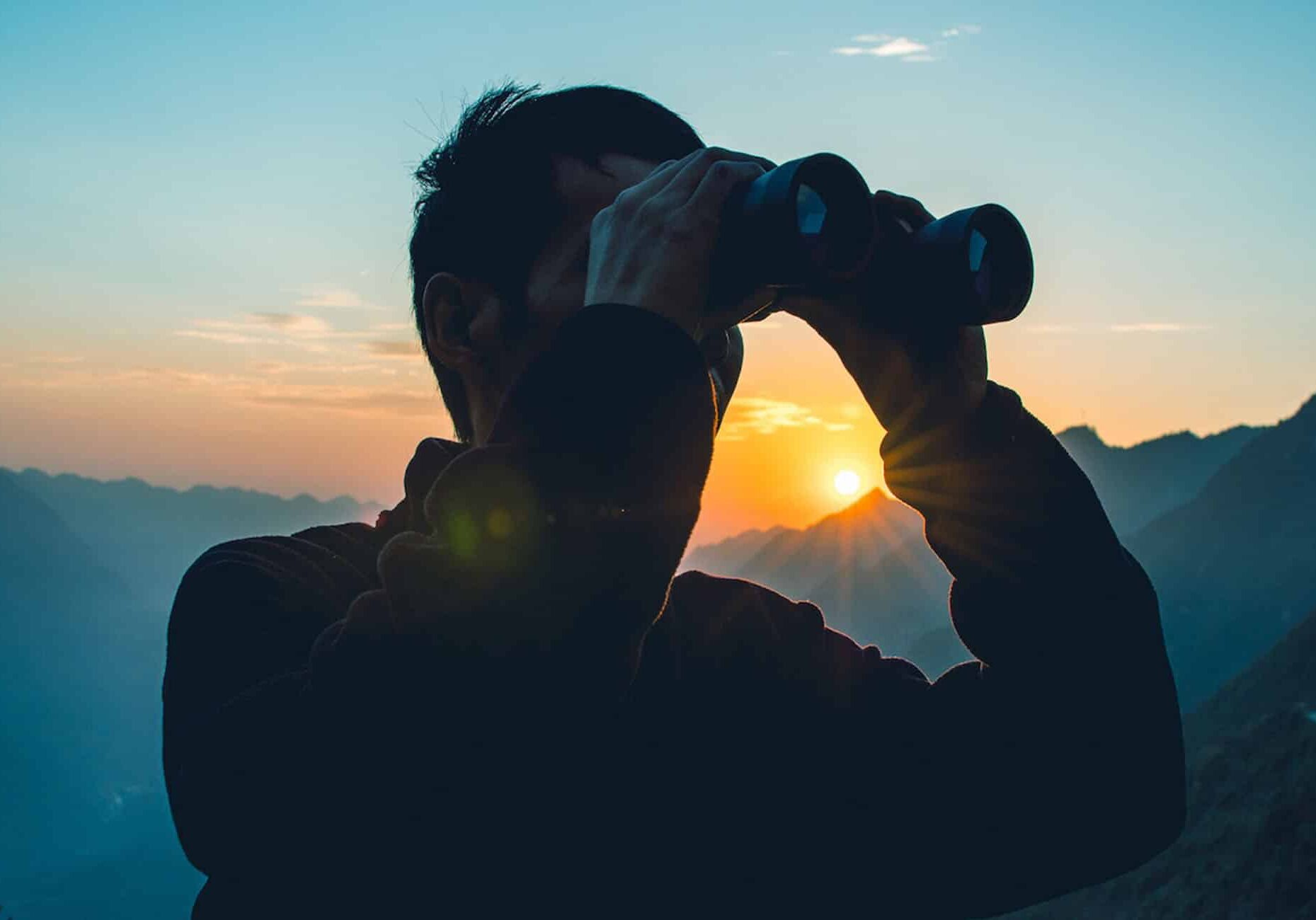 Man searching in binoculars with sunset behind him
